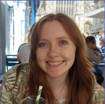 A smiling woman with long, straight red hair sits in front of a window with blue and white trim. She has fair skin, light blue eyes, and is wearing a green top with a floral pattern. She is holding a clear glass with a green straw, suggesting she is in a café or restaurant. The blurred background indicates a busy setting, possibly with other patrons, and part of a historic-looking stone building, likely a part of the café's street-side architecture, can be seen through the window behind her."