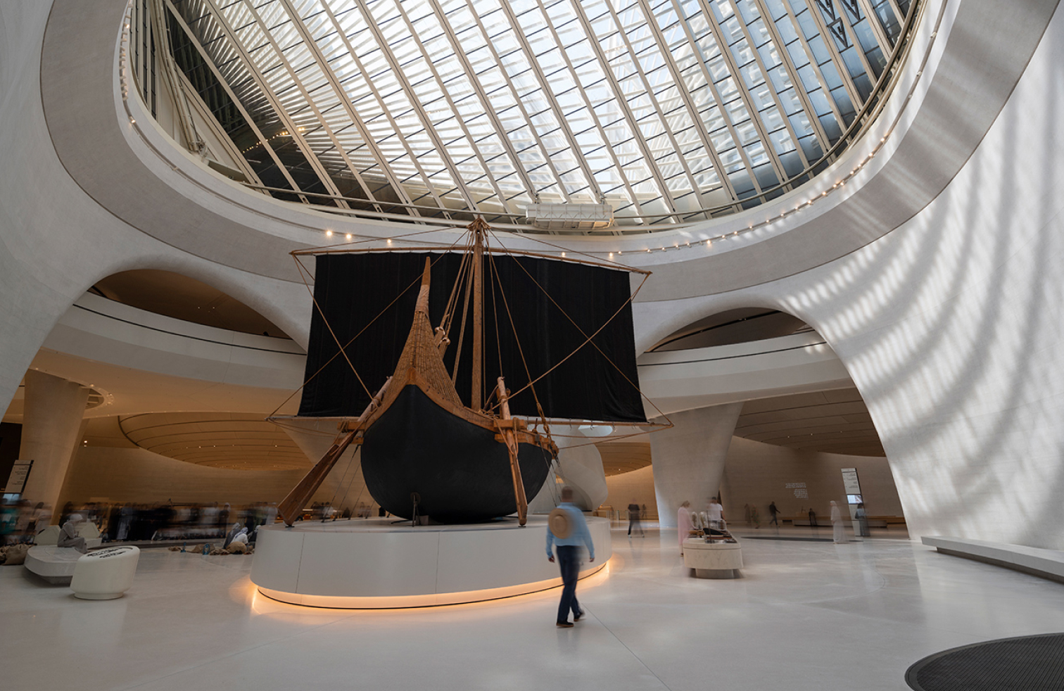 A traditional wooden sailboat with black sails displayed in a modern, white architectural gallery beneath a large skylight dome.