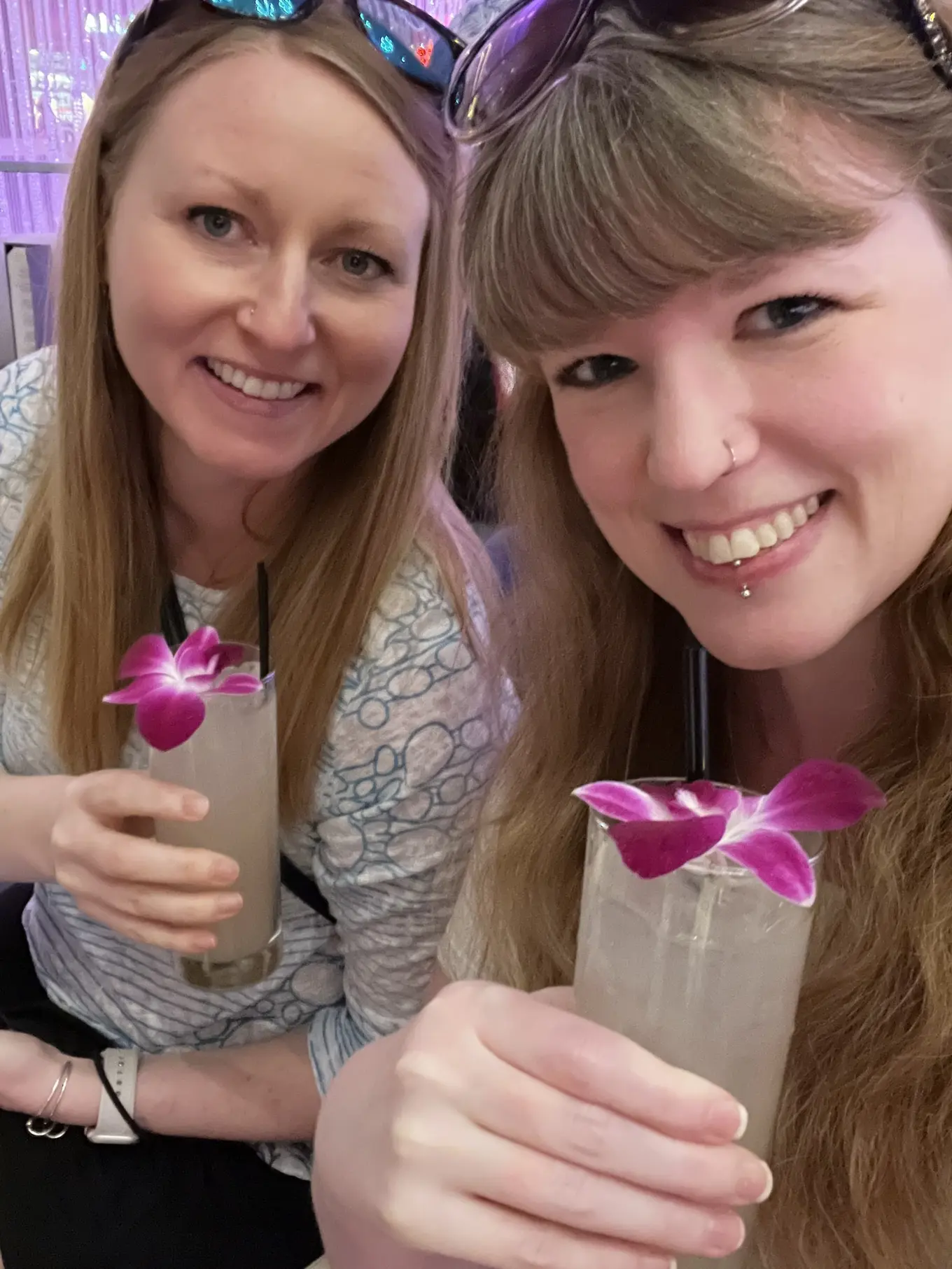 Two women smiling and holding drinks with purple orchid flowers as garnishes. They are both wearing sunglasses on their heads and enjoying a relaxed moment together, likely in a social or celebratory setting. The atmosphere appears casual and friendly.