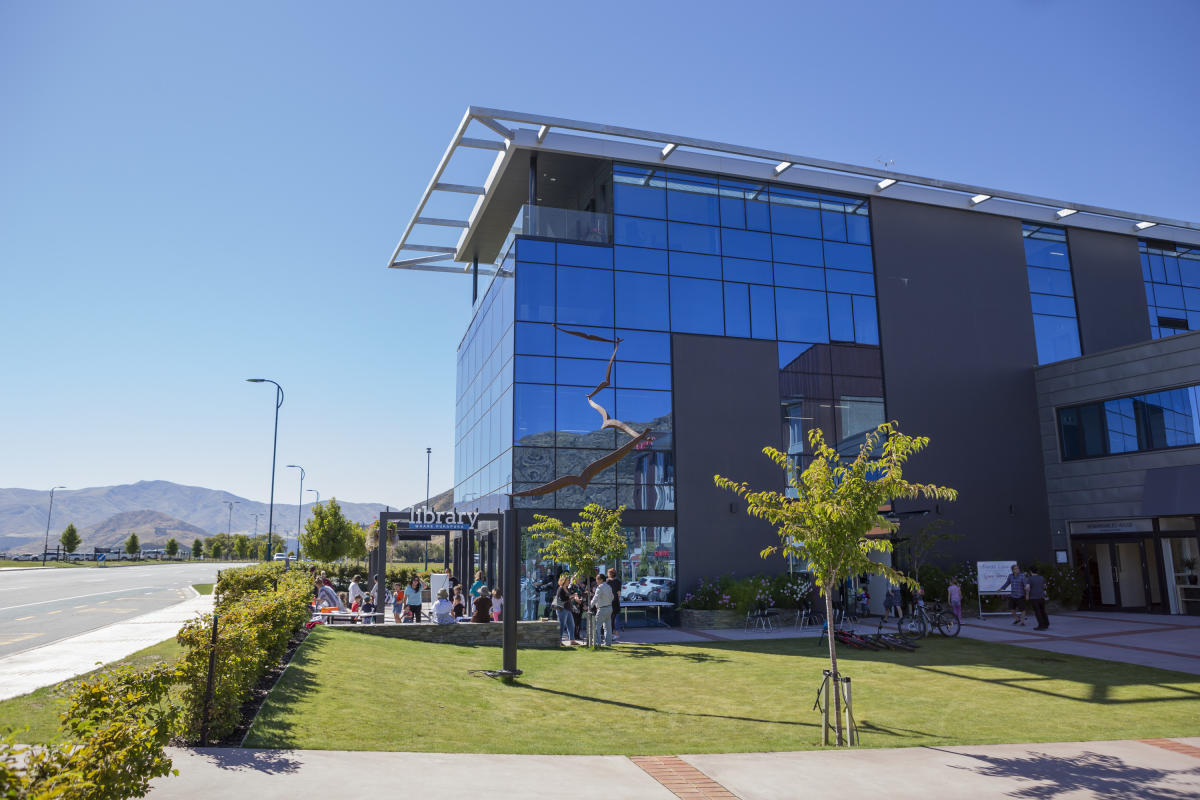 A wide exterior shot of a modern library building, featuring a blue glass facade, a manicured green lawn, and mountains in the background.