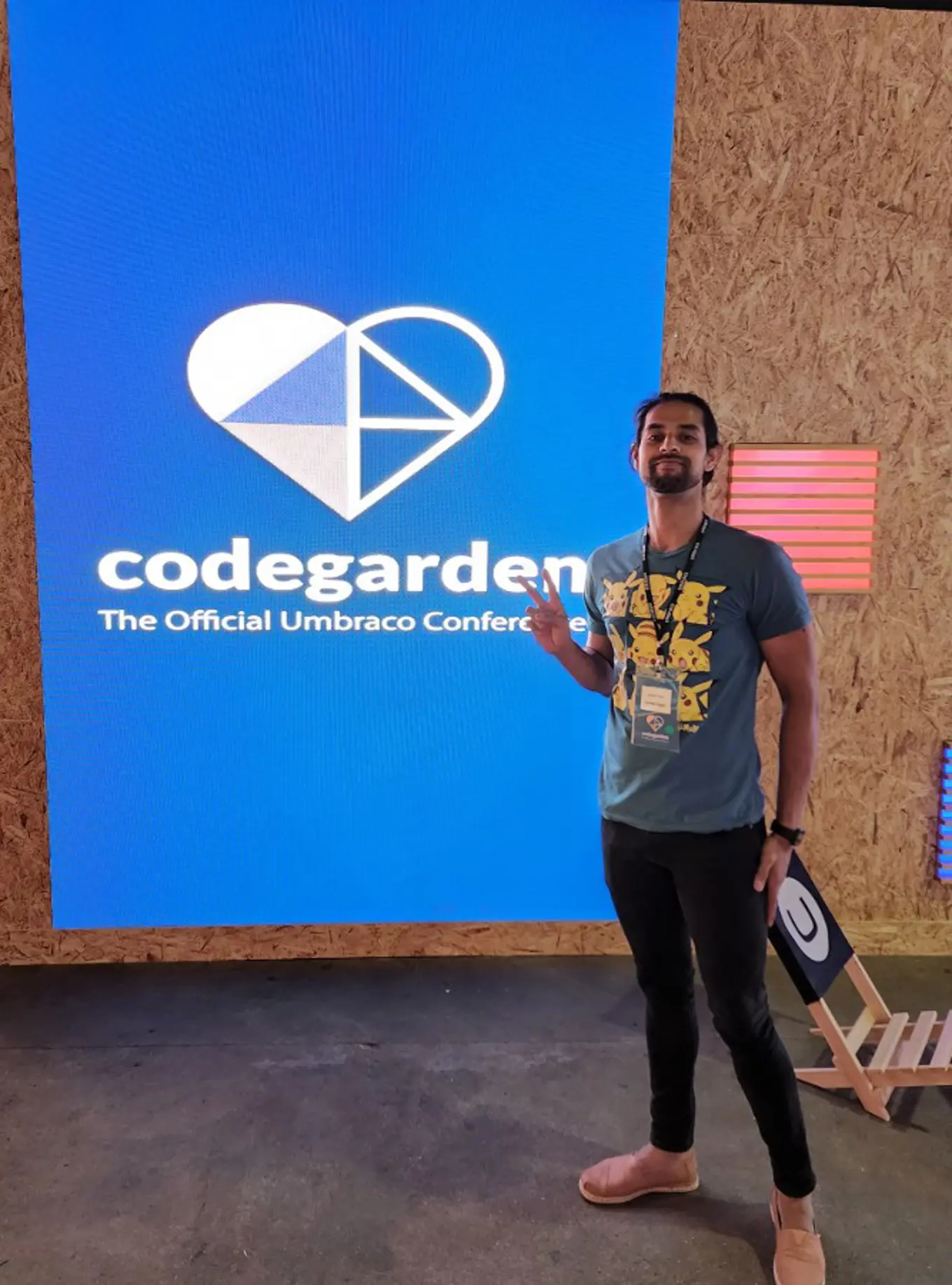 Javed stands in front of the led screen with the codegarden logo. He is proudly wearing a Pikachu t-shirt and giving the peace sign.