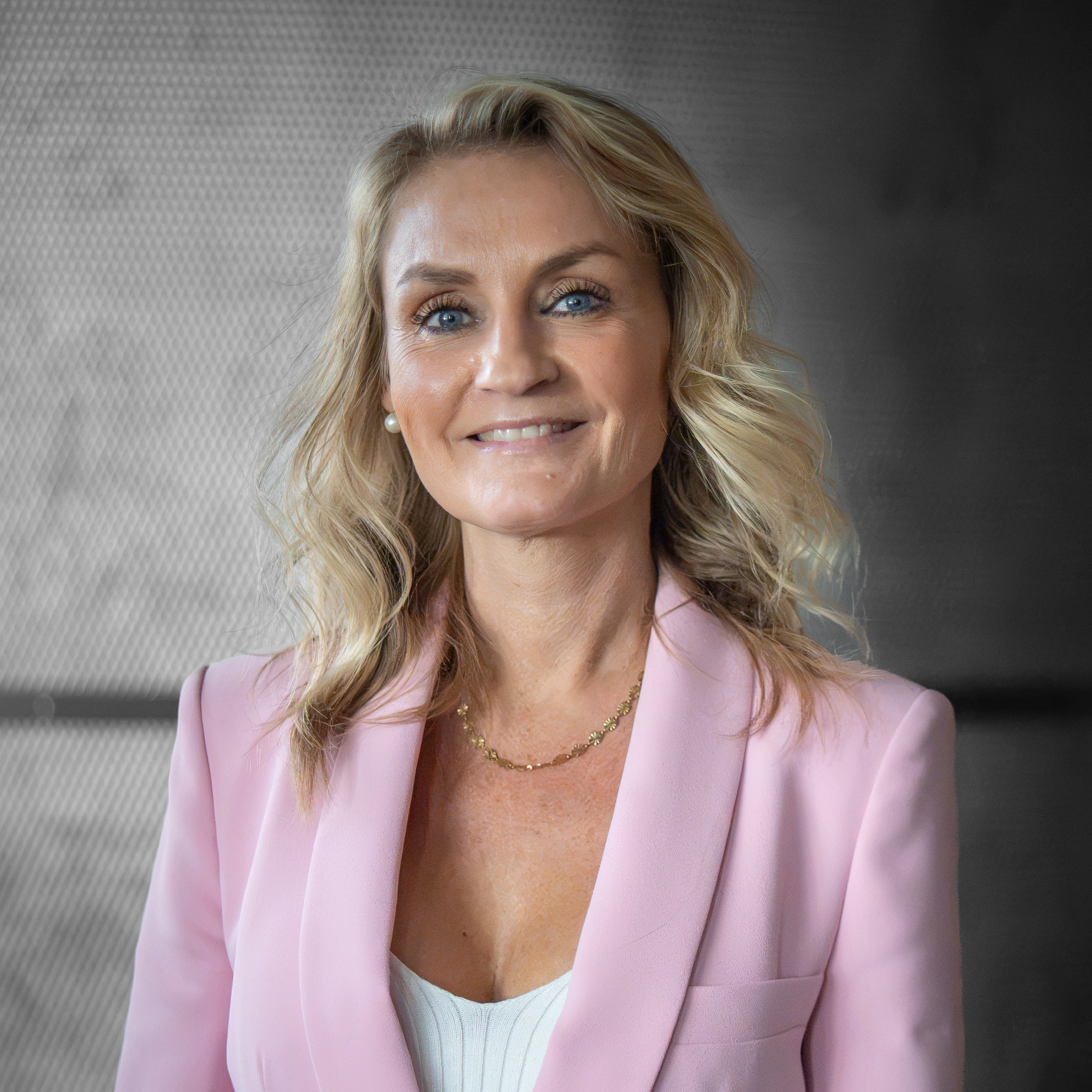 Smiling blonde woman in a light pink blazer portrait against a textured gray background.
