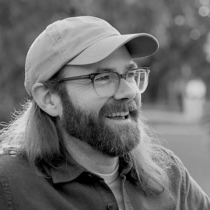 Profile Picture of Allen Smith, smiling and looking into the distance. Wearing a baseball cap and glasses, sporting a beard. Photo in black and white