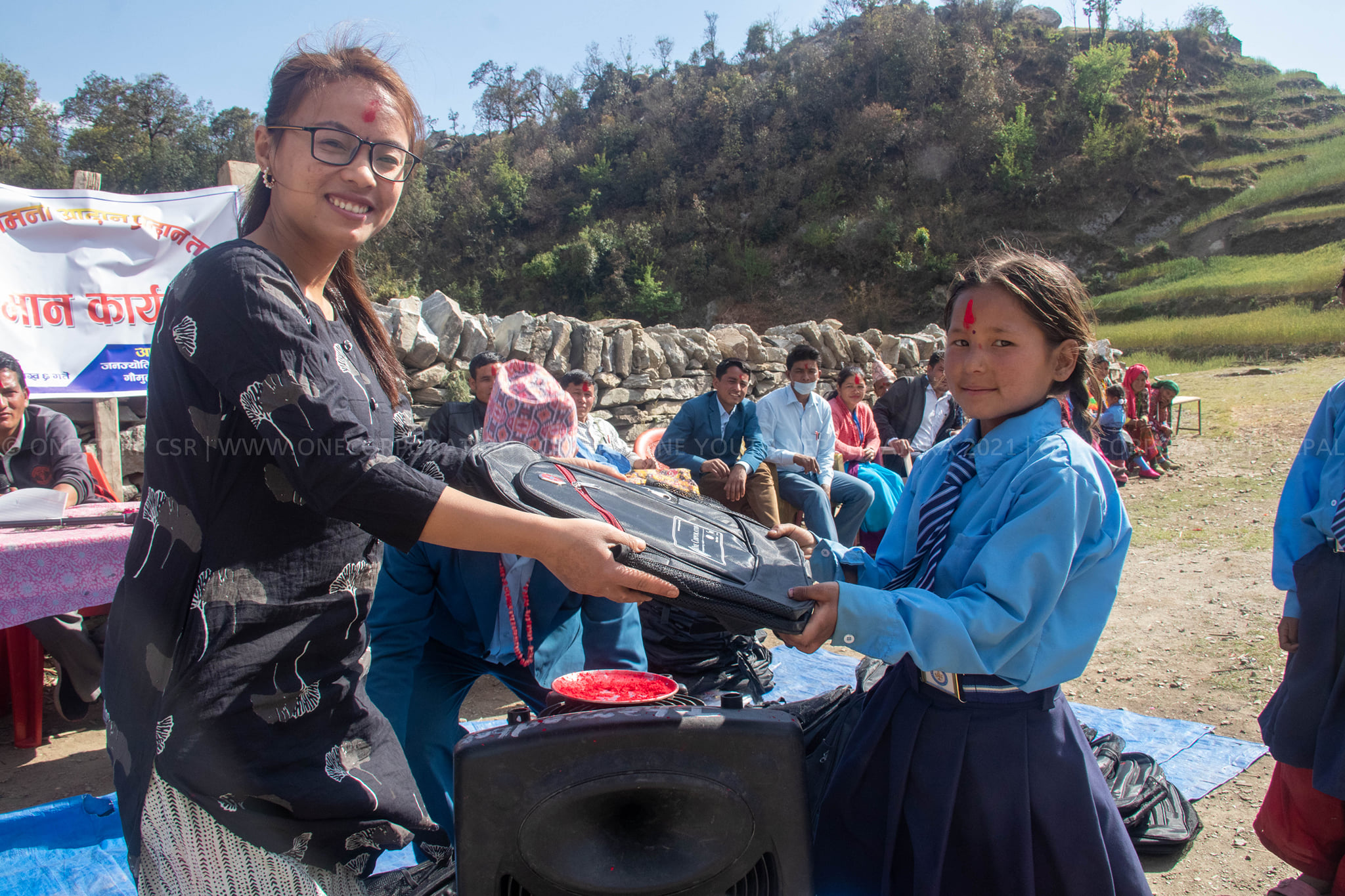 A woman wearing glasses hands a black school backpack to a young girl in a blue uniform during an outdoor community distribution event.