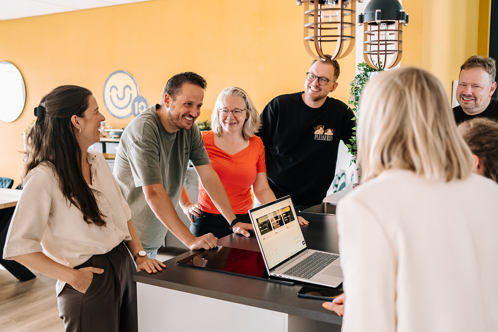 Team of six smiling in an office meeting around a laptop on a counter with coffee-bar decor in back