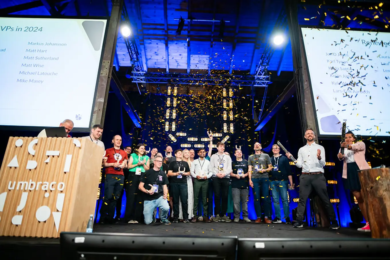 A group of people standing on a stage under confetti, celebrating with awards, while two large screens display text in the background.