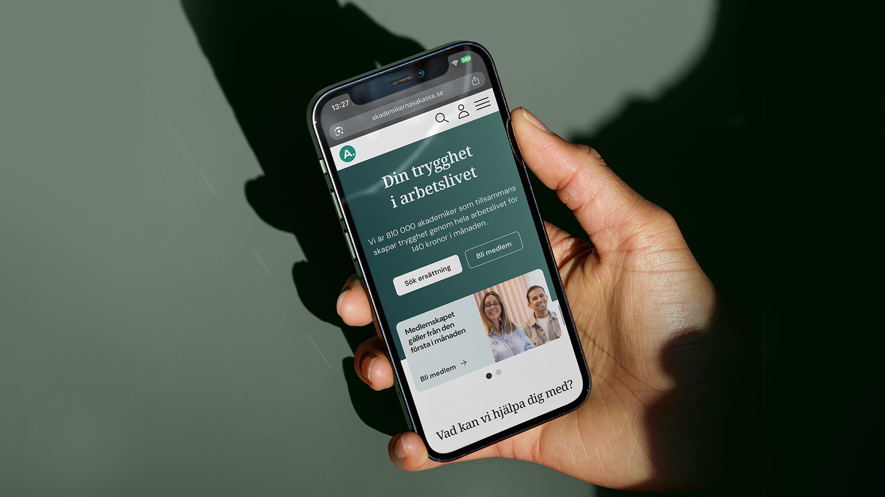 A hand holding a smartphone displaying the mobile-responsive layout of the Akademikernas a-kassa homepage.