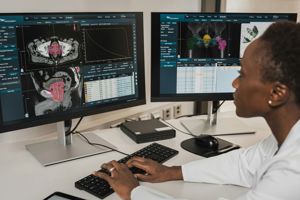 A healthcare professional working at a desk with two computer monitors displaying medical imaging scans and specialized radiology software.