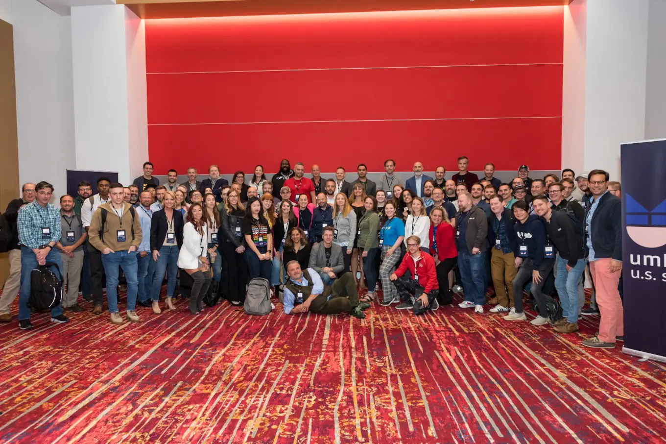 A group of 100 people smiling in a group in a room with red rug and carpet