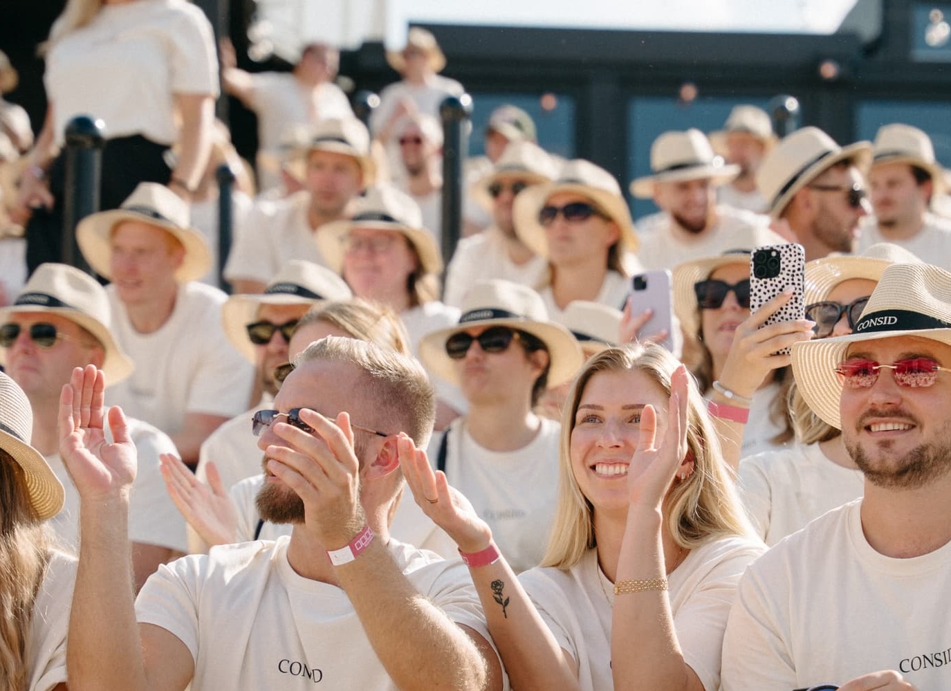 Crowd at an outdoor event, many people in white hats and shirts, smiling and waving with phones in hand