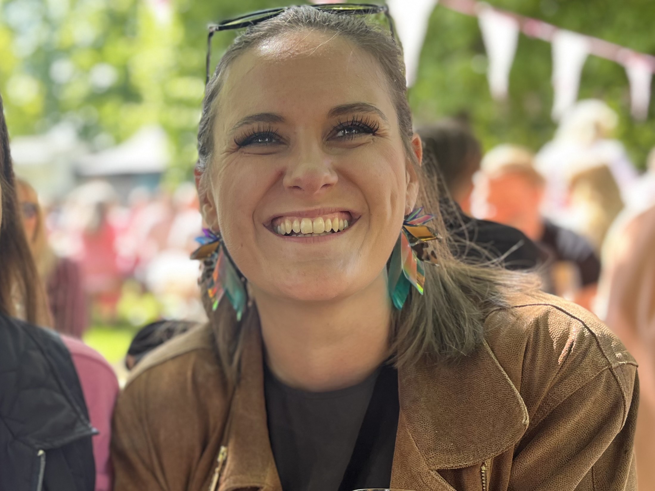 A close-up of a person smiling brightly while wearing colorful, feather-like earrings and a brown jacket. The background is a soft-focus outdoor setting with sunlight filtering through trees and other people visible in the distance.