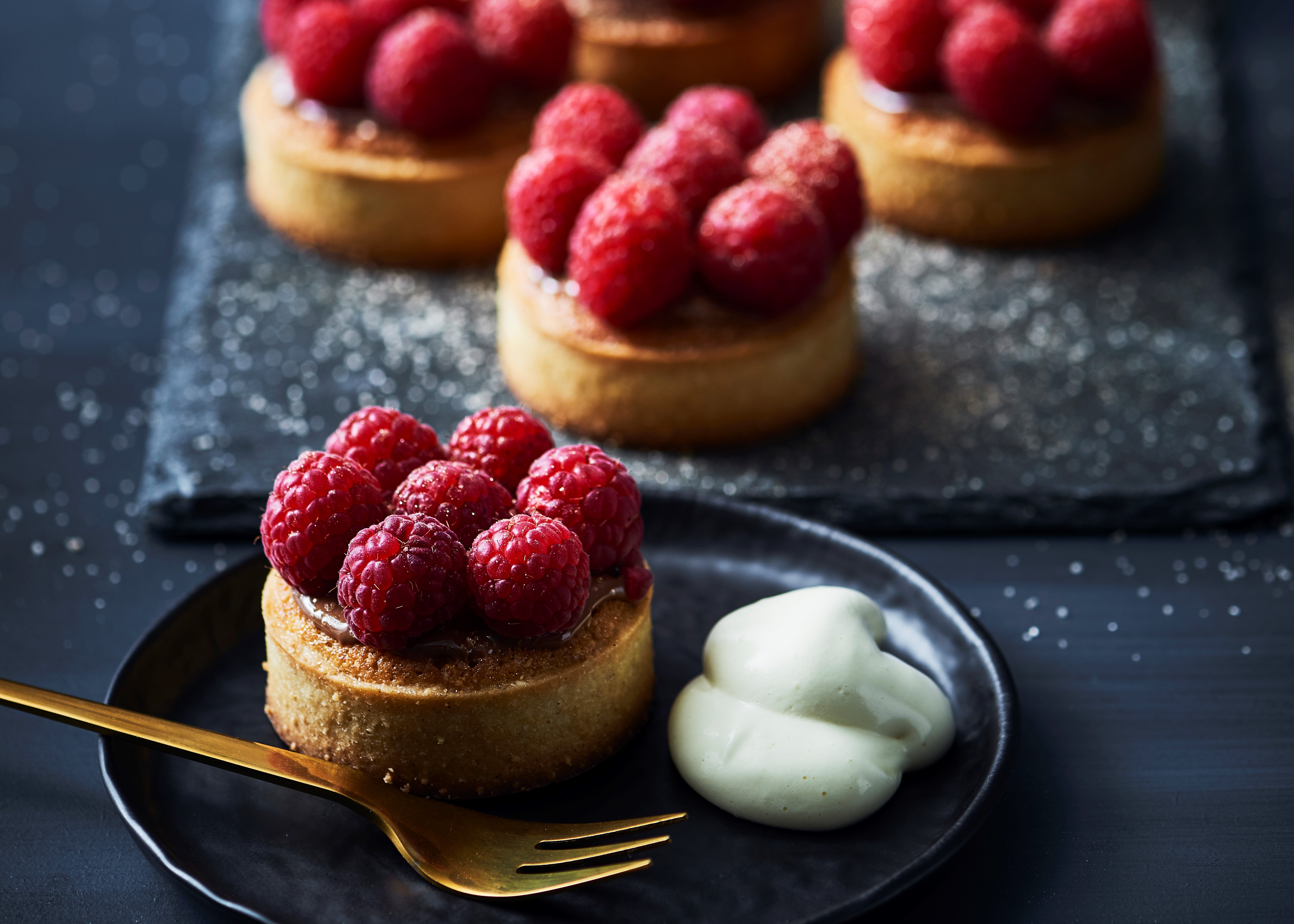 Several small, round pastry tarts topped with fresh raspberries and a dollop of cream on a dark plate.