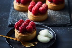 Several small, round pastry tarts topped with fresh raspberries and a dollop of cream on a dark plate.
