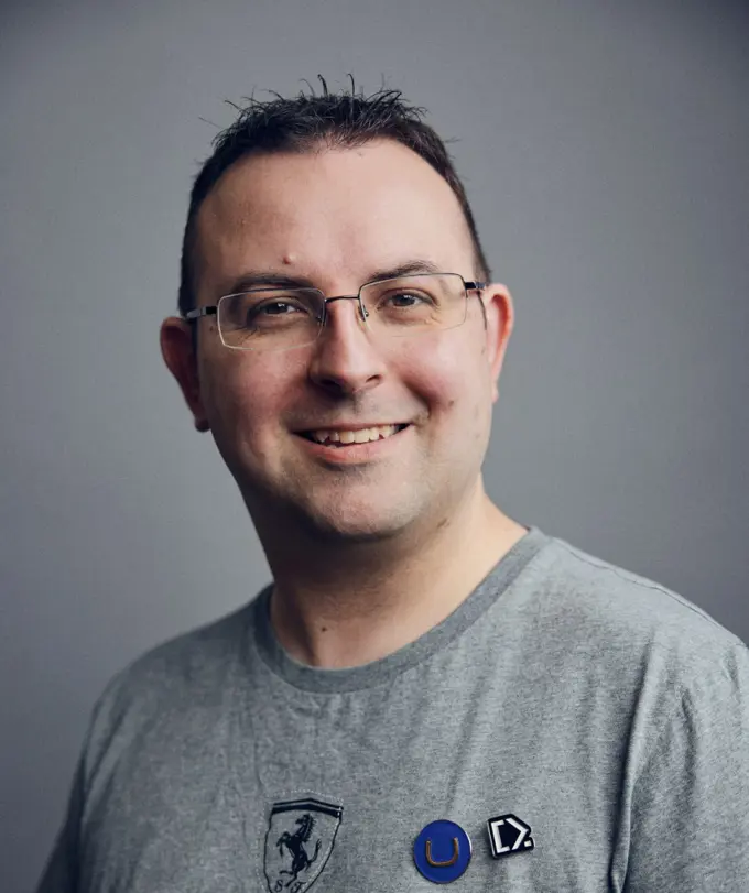 A professional headshot of a smiling man wearing a grey t-shirt with Umbraco and Codecabin pins, standing against a grey background.