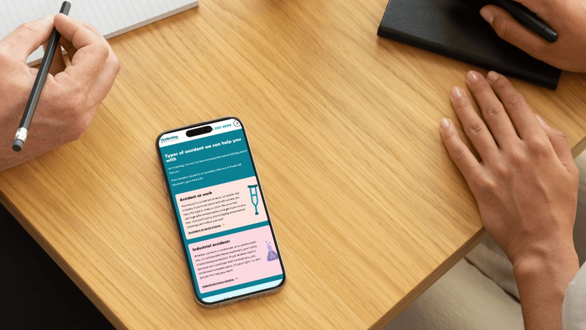 A smartphone on a wooden desk displaying the Underdog website, showing claim categories for "Accident at work" and "Industrial accidents."