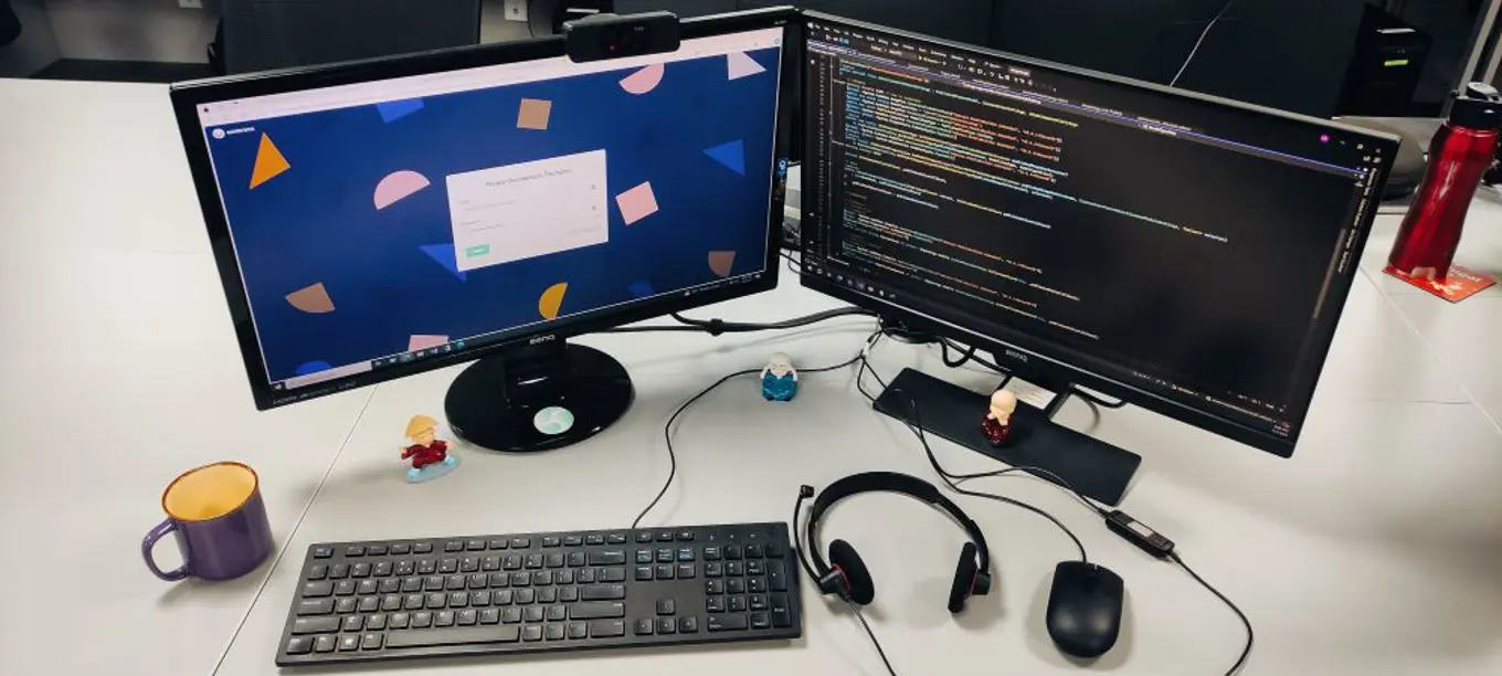 Image of Dhanesh's desk at work, showing two monitors displaying code and the Umbraco back office log in screen.