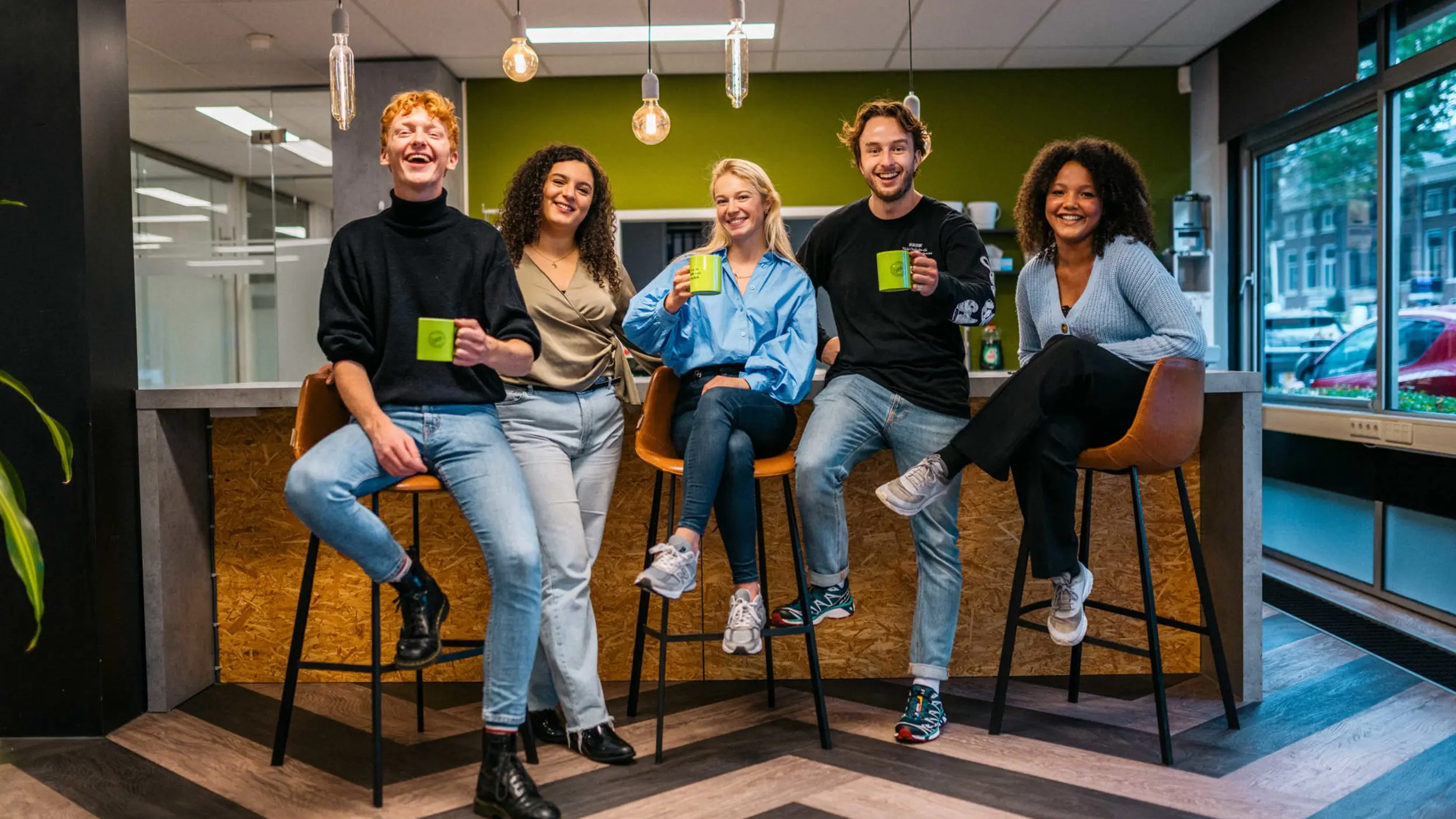 A group of five smiling young adults holding matching lime-green mugs in a modern office breakroom.