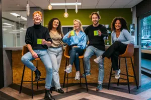 A group of five smiling young adults holding matching lime-green mugs in a modern office breakroom.