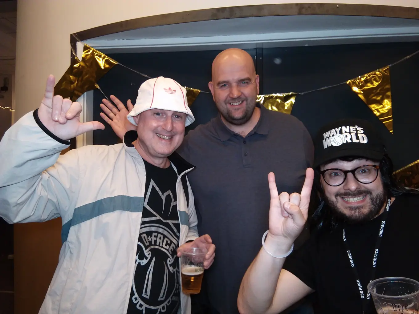 A photo of Rick Butterfield with Dean Leigh and Paul Seal at the Codegarden 24 pre-party held at Umbraco HQ. The photo is taken against a dark painted door, with shiny gold bunting in the background and they are wearing 90's themed clothing.