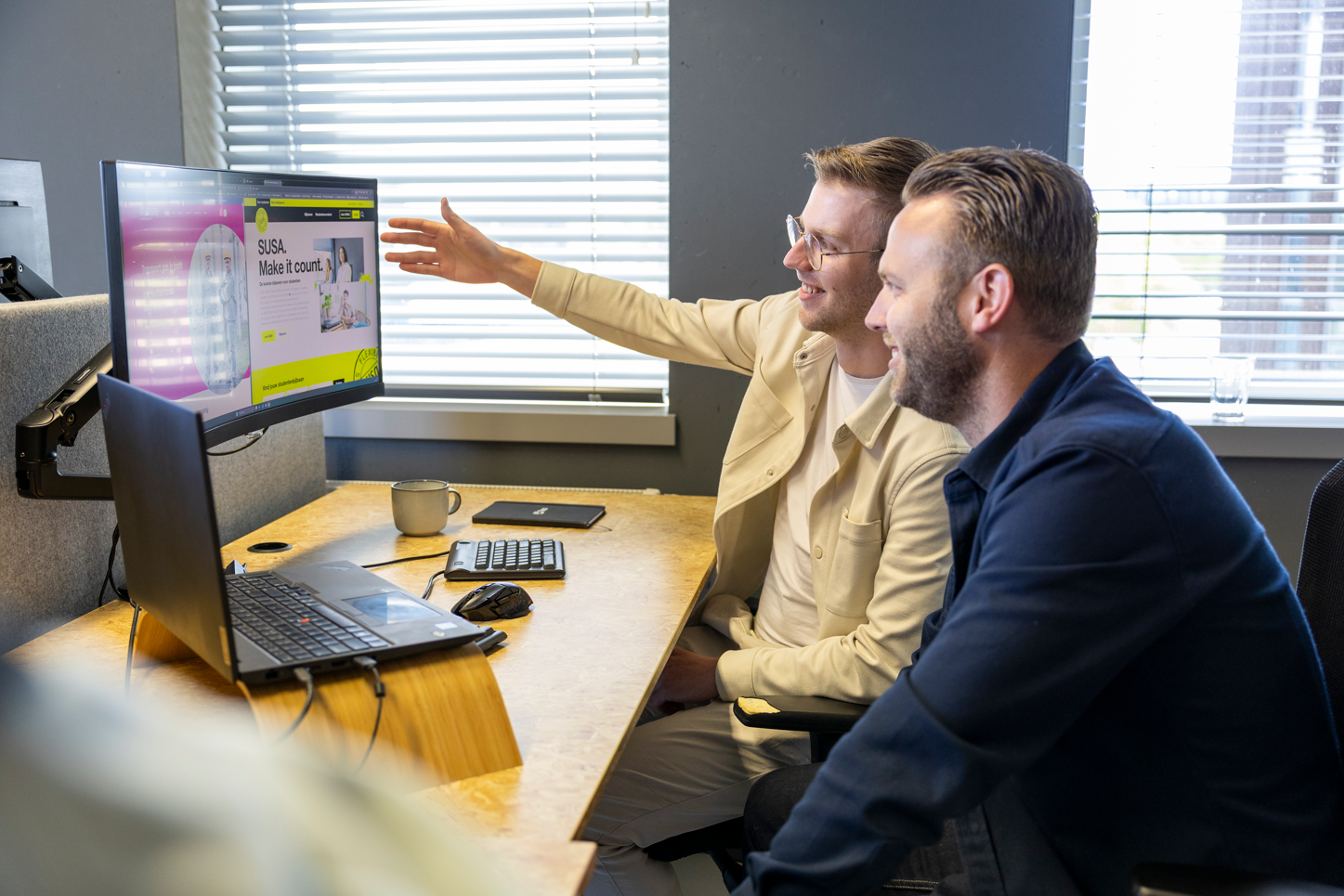 Two men in an office setting discussing the SUSA website displayed on a large desktop monitor.