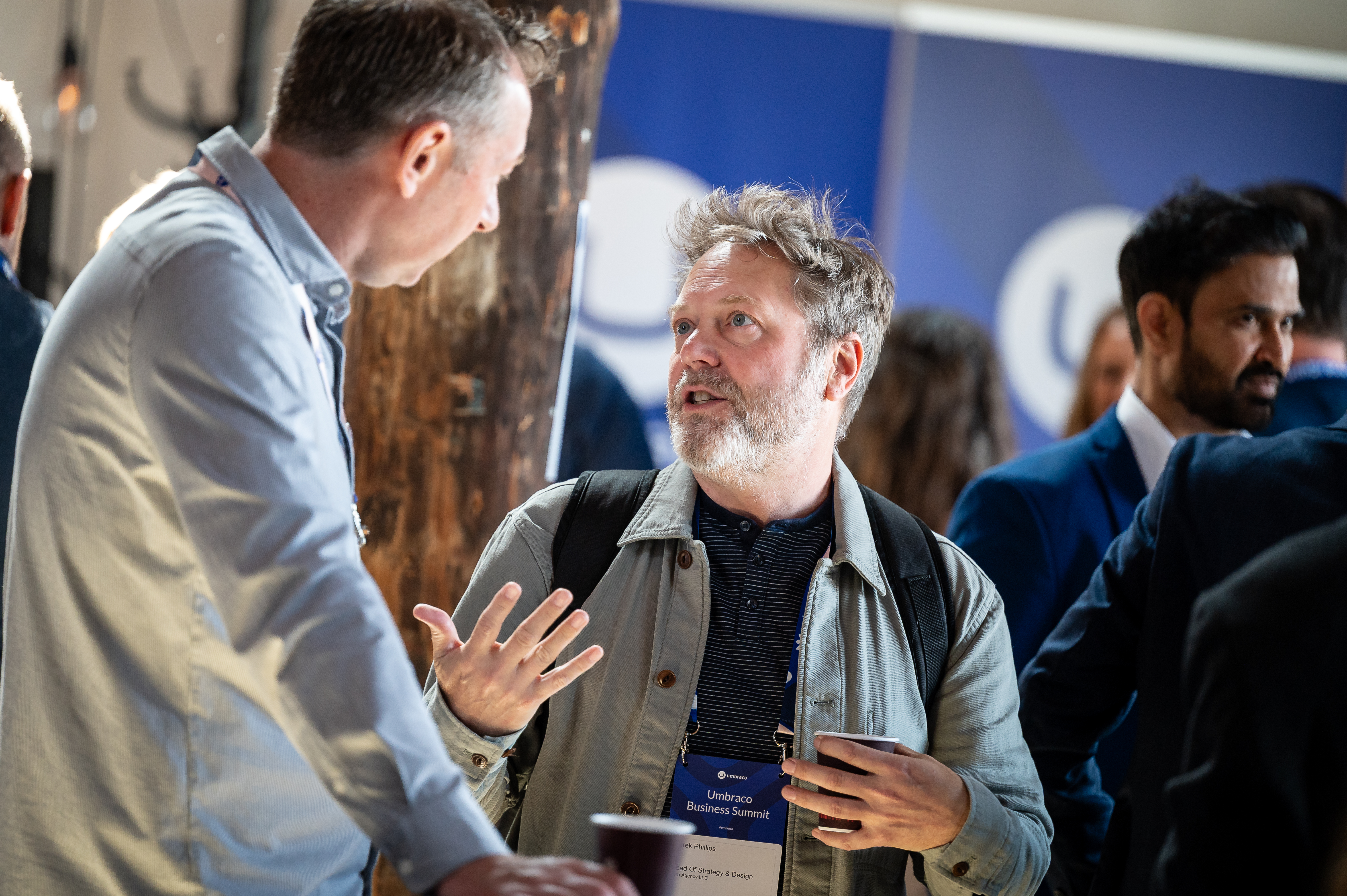 Two men in conversation at a business event, one gesturing, with a blue backdrop behind them.