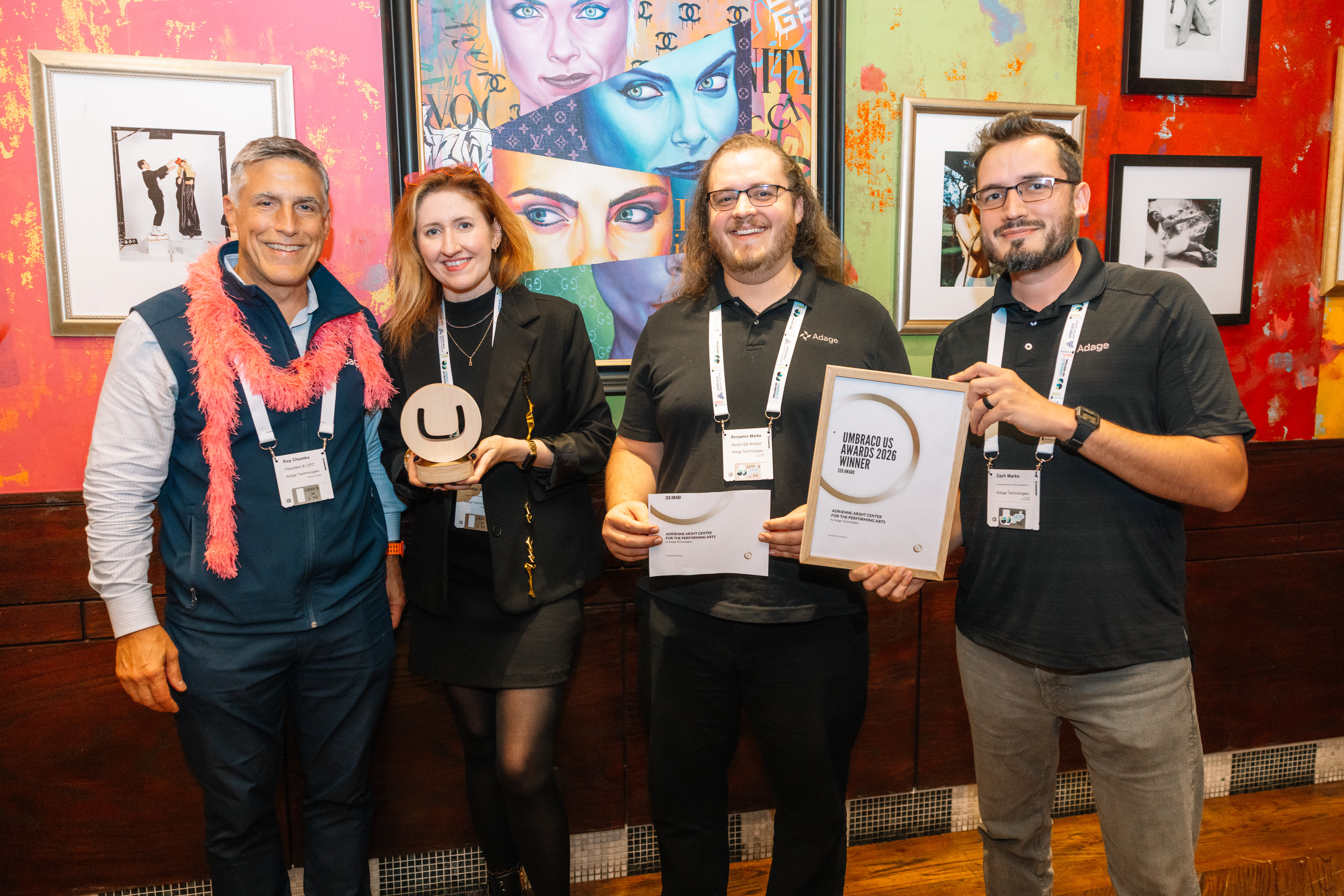 Four people at an event holding awards and certificates in front of colorful wall art and framed photos