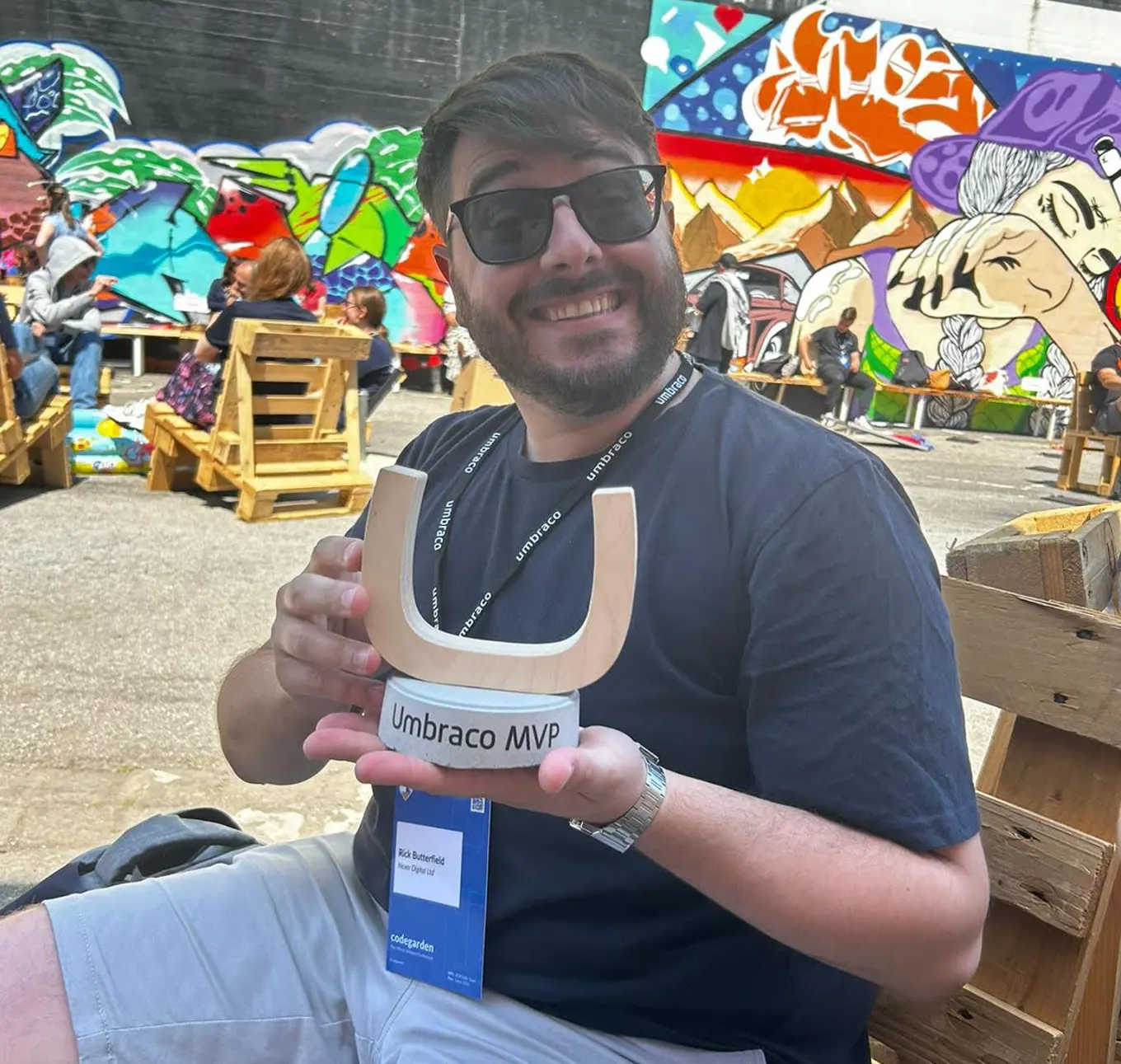 Rick Butterfield, beaming with pride, holding his freshly won Umbraco MVP Award, while at Codegarden 2023. He is sat outside on a wooden pallet chair, wearing light grey shorts and a dark blue t-shirt and sunglasses, in front of a vibrantly colourful graffiti wall.