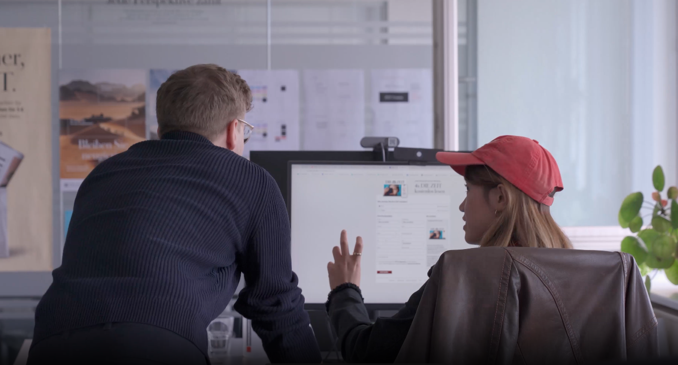Two people in a modern office viewing a computer screen with a desk display of documents and charts