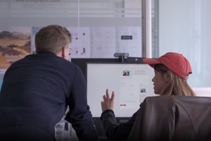 Two people in a modern office viewing a computer screen with a desk display of documents and charts