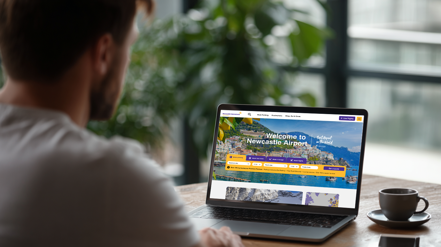 A man using a laptop to browse the "Welcome to Newcastle Airport" website at a wooden table.