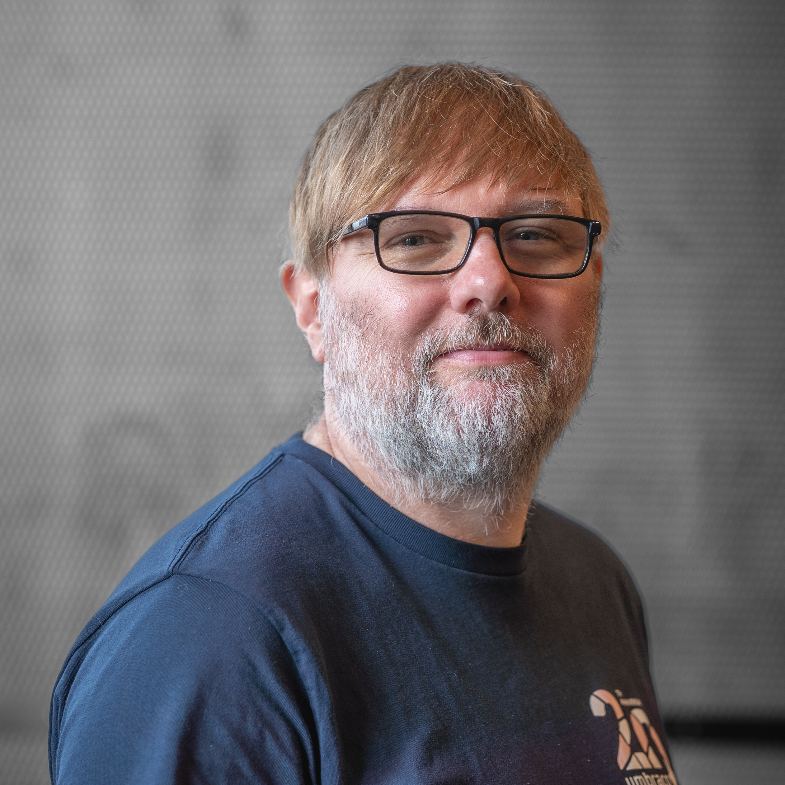 A portrait of a man with blonde hair, a grey beard, and glasses wearing a dark blue T-shirt. He is looking toward the camera with a neutral, pleasant expression against a textured grey background.
