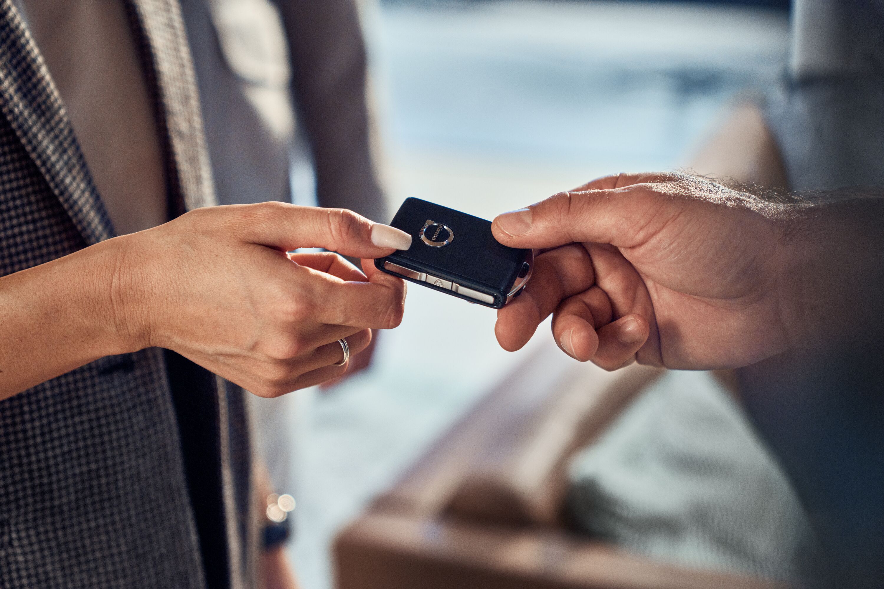 A close-up shot of a person in a grey patterned jacket handing a black Volvo key fob to another person.