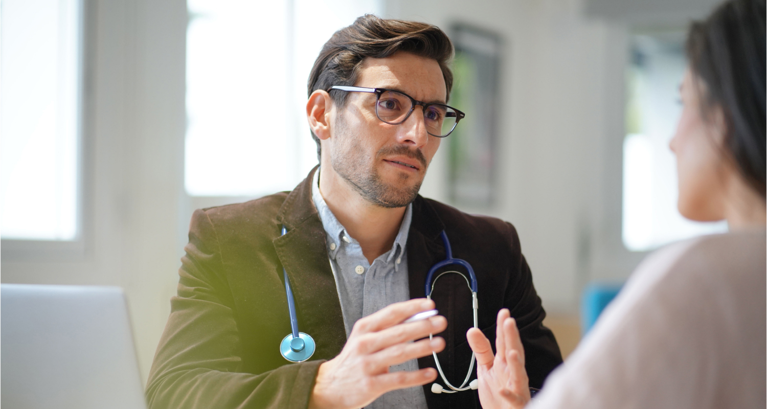 A close-up of a male doctor wearing glasses and a stethoscope, engaged in a focused conversation with a patient in a bright medical office.