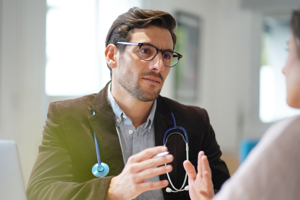 A close-up of a male doctor wearing glasses and a stethoscope, engaged in a focused conversation with a patient in a bright medical office.