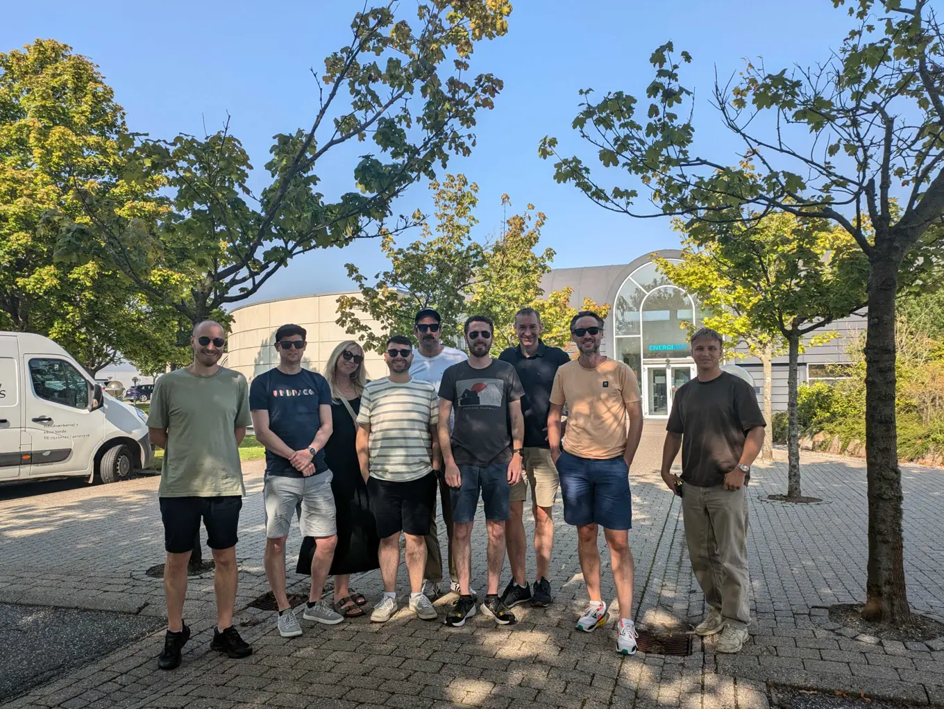 A group photo of the Sustainability team, made up of men and women, taken on a sunny day, with blue sky and green trees in the background, as they visit EnergiNet in Denmark.