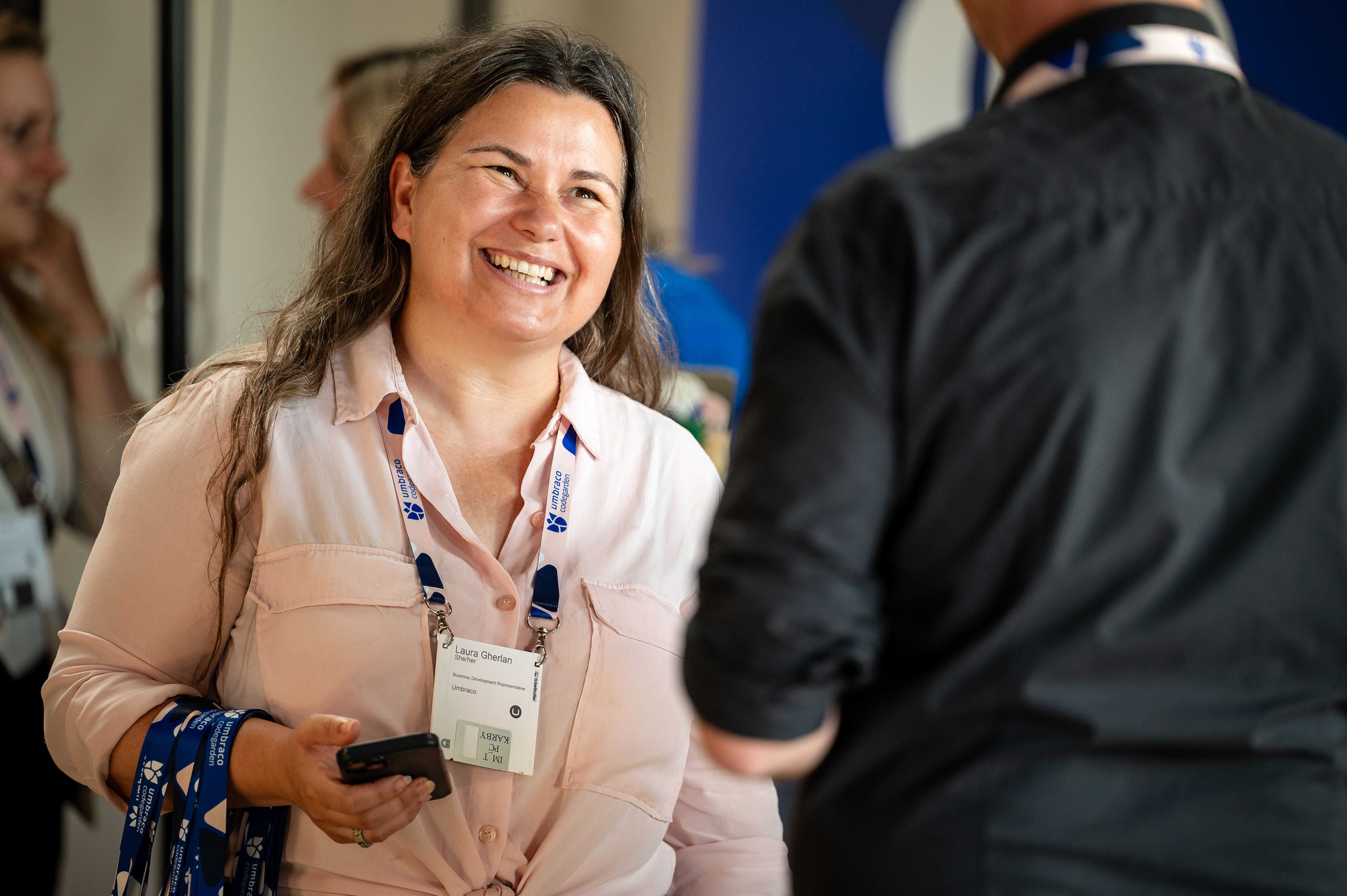 Smiling woman in a light shirt holding a phone, wearing an ID badge at an event conversation.