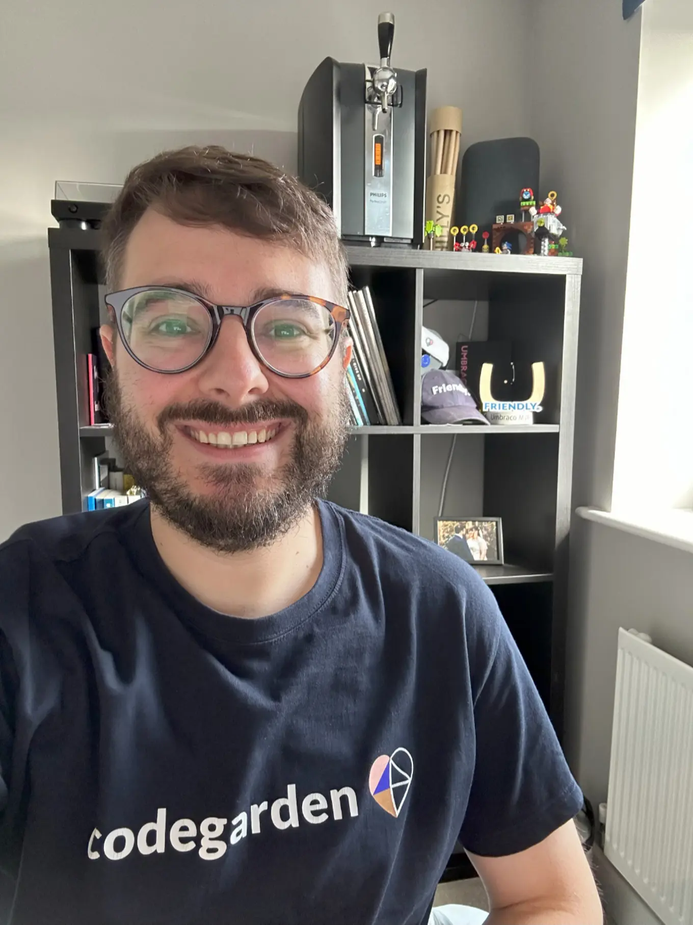 A picture of Rick Butterfield wearing an dark blue Umbraco logo t-shirt, smiling in front of a set of shelves which house all of his Umbraco memorabilia.