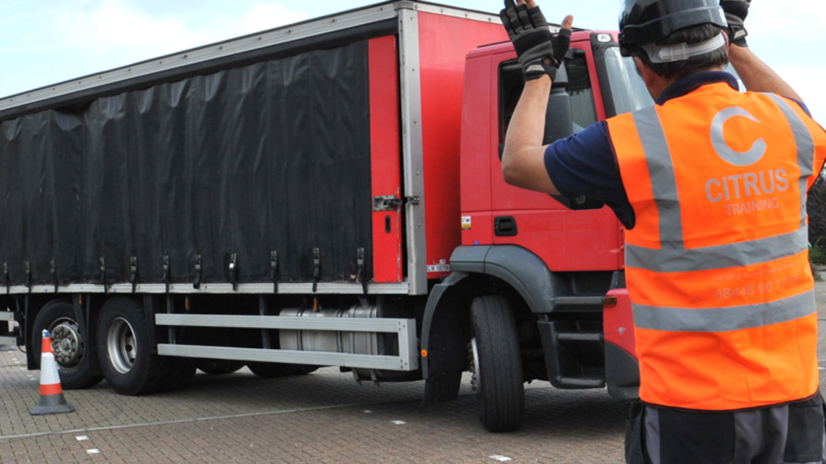 A close-up, real-life photo of a person wearing an orange high-visibility Citrus Training vest and helmet as they guide a large red commercial truck.