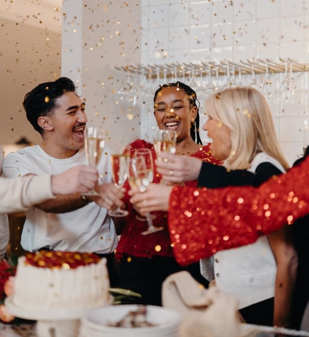 Group of friends laughing and clinking champagne glasses in front of a small white cake, with confetti falling.