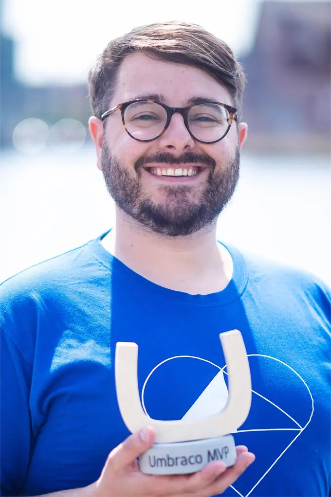 Profile Picture of Rick Butterfield. A smiling white man with dark hair and a beard, wearing glasses, a blue Umbraco t-shirt and holding an award. He is standing in front of a body of water, with buildings beyond, which are fussy in the background, on a bright, sunny day.