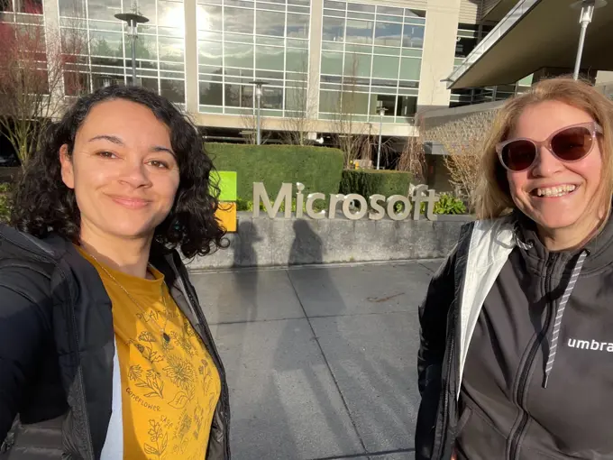 Two women taking a selfie in front of the Microsoft headquarters sign on a sunny day. Both are smiling broadly; one wears a yellow t-shirt and a black jacket, the other a black jacket, Umbraco hoodie, and sunglasses.