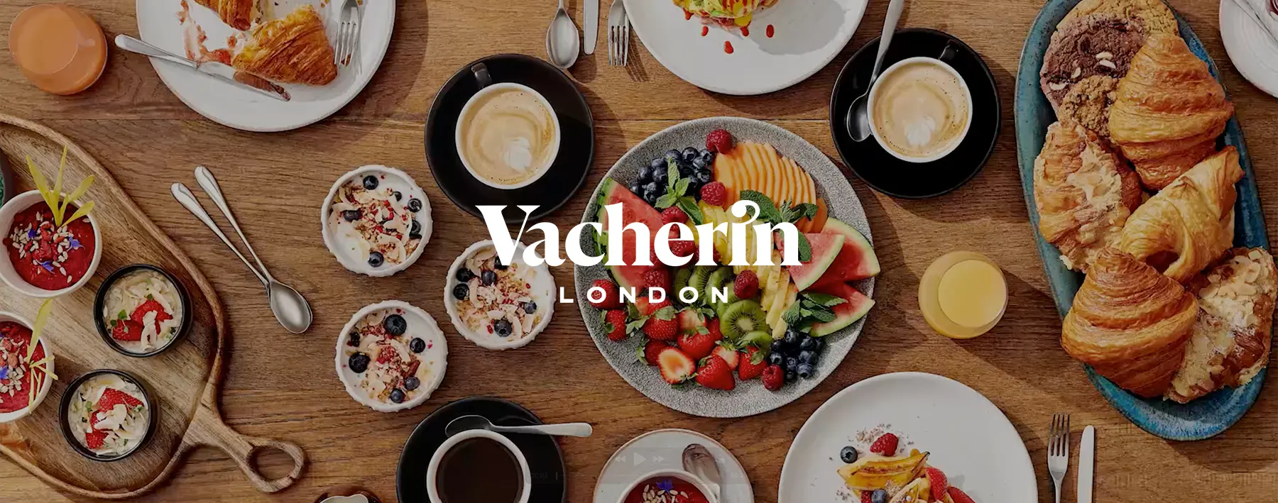 A top-down view of a large wooden table spread with various breakfast foods, including fruit platters, croissants, yogurt bowls, and coffee, with the "Vacherin London" logo centered in white.