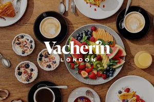 A top-down view of a large wooden table spread with various breakfast foods, including fruit platters, croissants, yogurt bowls, and coffee, with the "Vacherin London" logo centered in white.