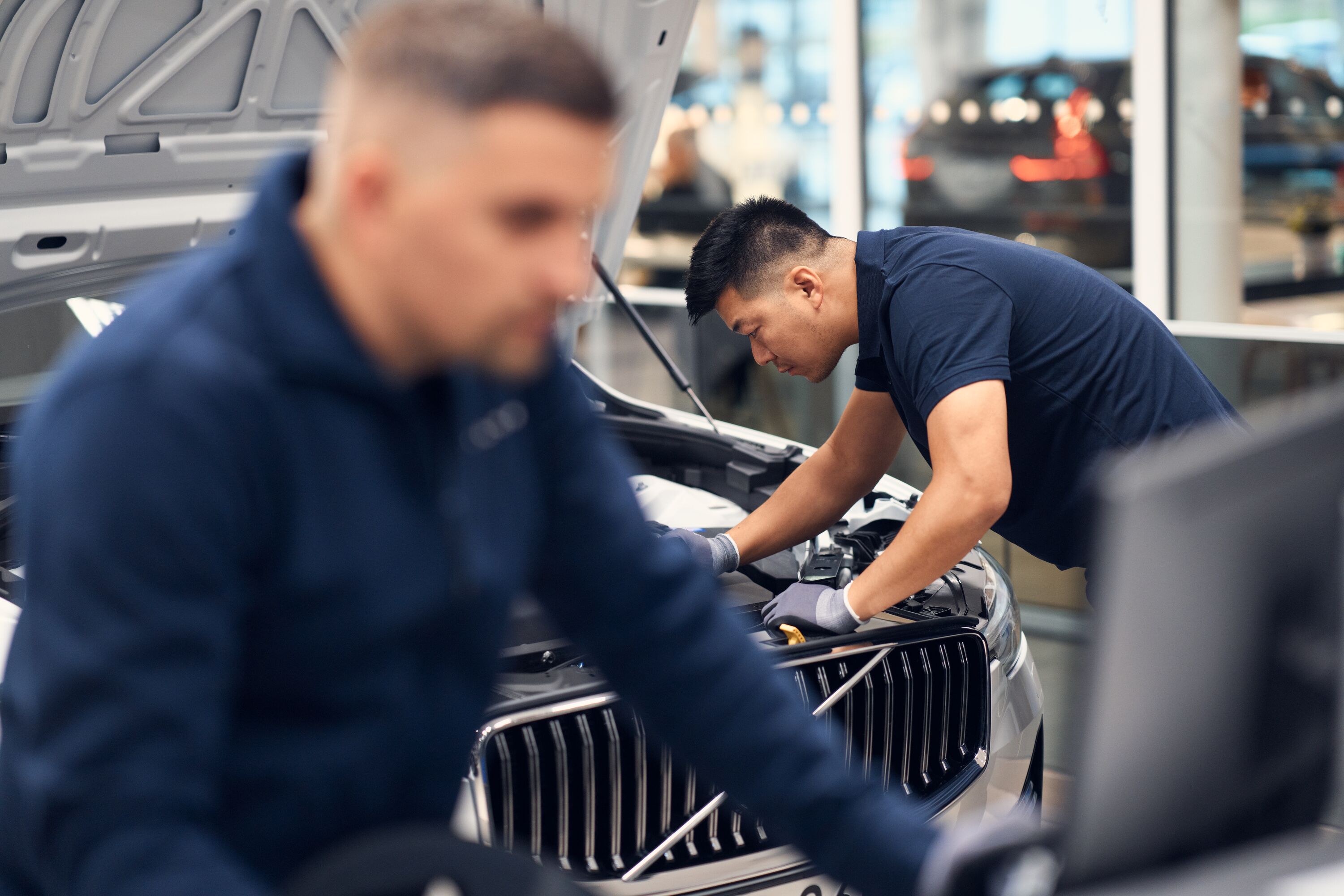 A mechanic in a dark blue polo shirt leans over the open hood of a silver Volvo to perform engine maintenance in a bright, modern service workshop.